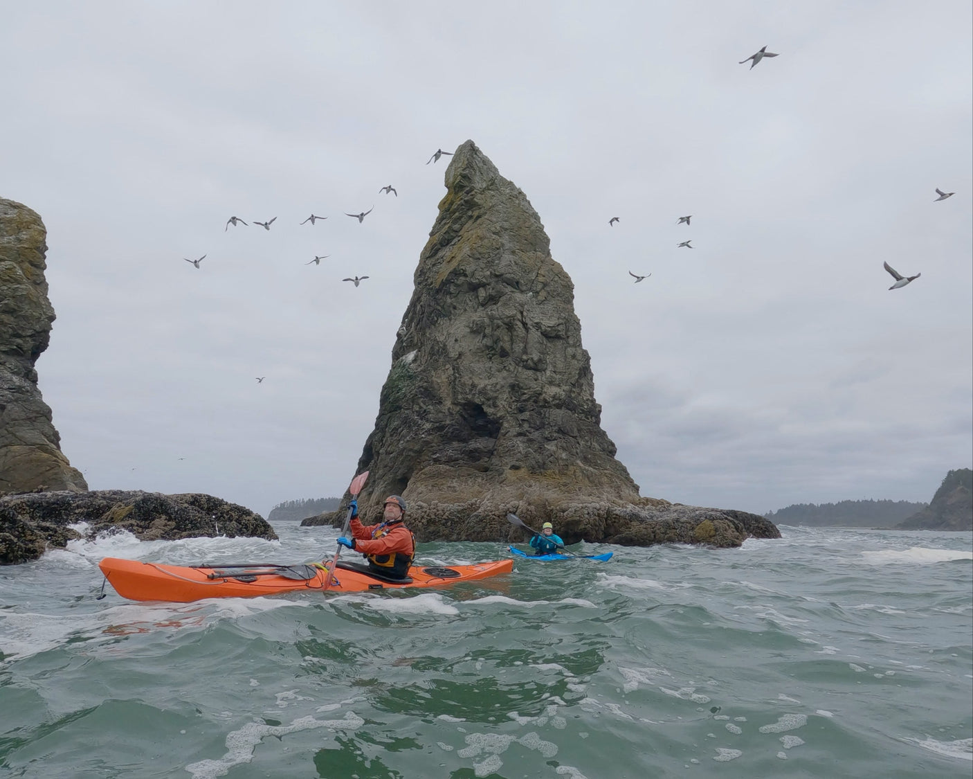 Two kayakers paddling near large rock formations with birds flying overhead.